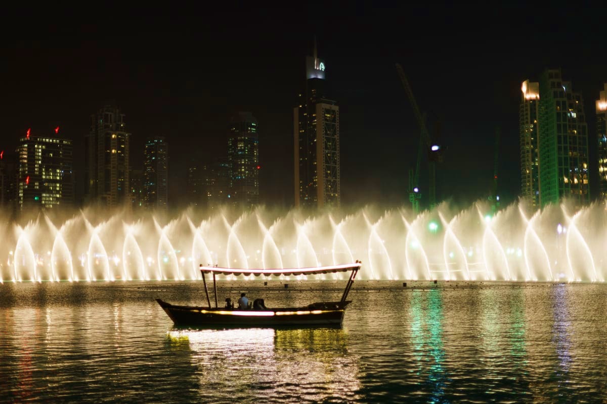 Illuminated fountain display at night with traditional dhow boat in foreground and Dubai city skyline with lit buildings in b