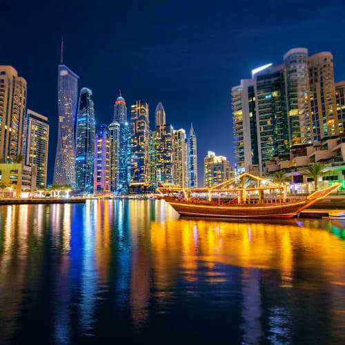 Dubai Marina skyline at night with illuminated skyscrapers and traditional wooden dhow boat on calm water