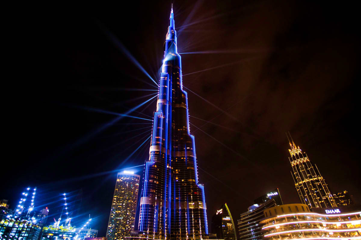 Dubai Grand Stopover Experience Burj Khalifa illuminated at night with blue and white lights against dark sky, surrounded by other lit buildings in Dubai sky