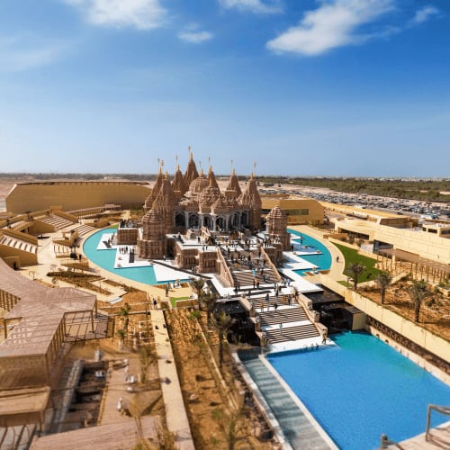 Aerial view of a large ornate temple with multiple spires surrounded by turquoise swimming pools and sandy desert landscape i