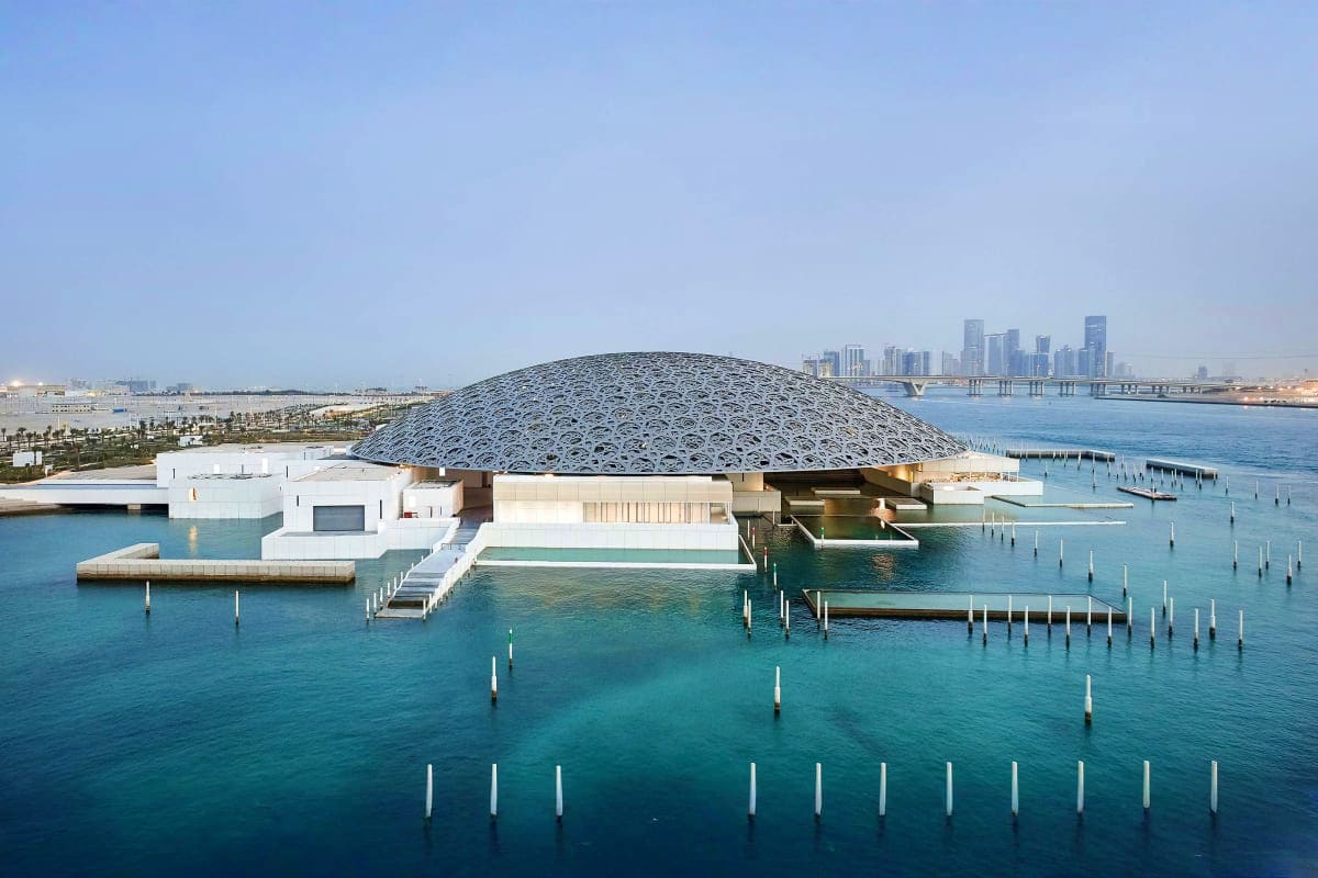 Louvre Abu Dhabi museum with distinctive dome on waterfront surrounded by turquoise water and mooring posts, with Dubai skyli