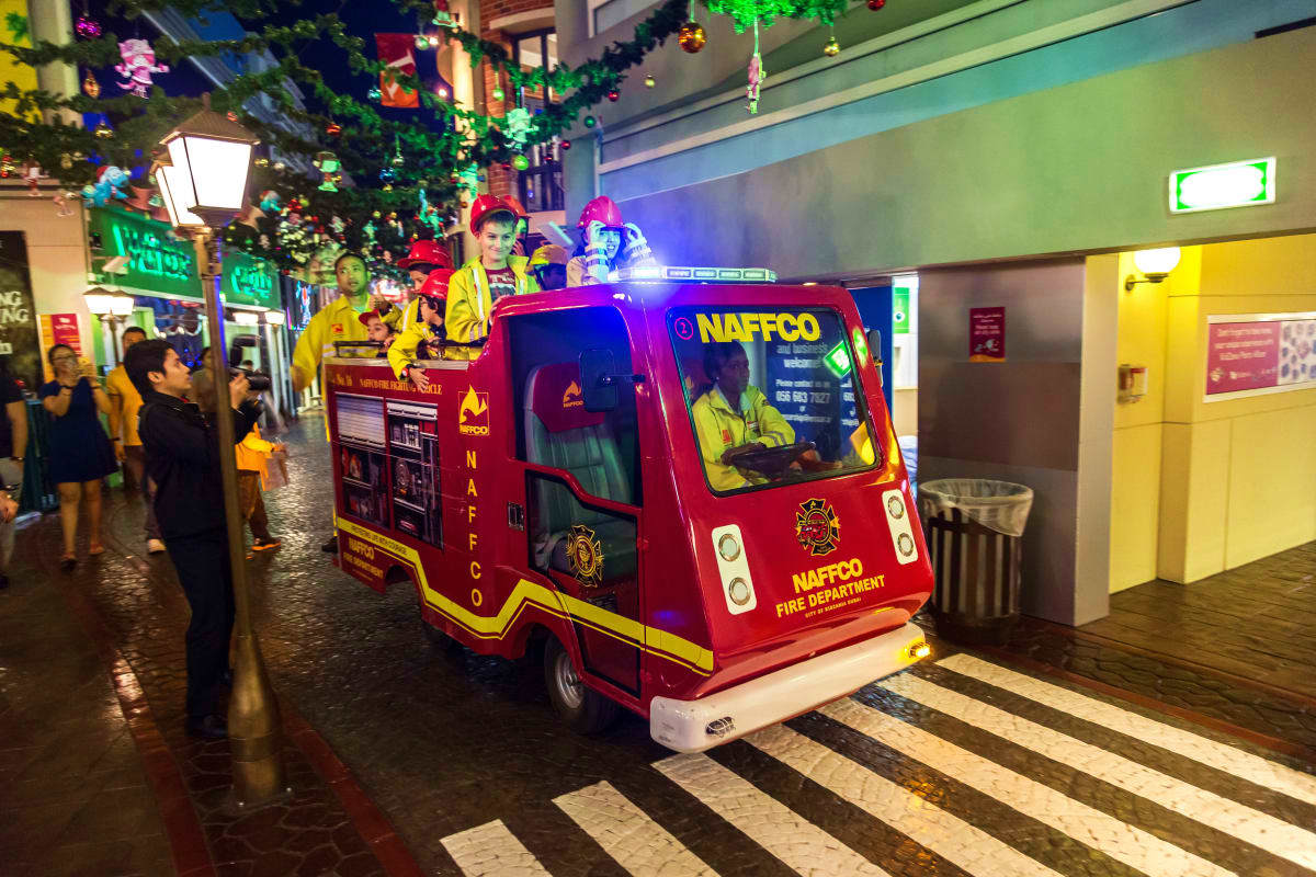 Red and yellow NAFFCO fire truck ride with children in festive costumes at indoor holiday market with colorful decorations