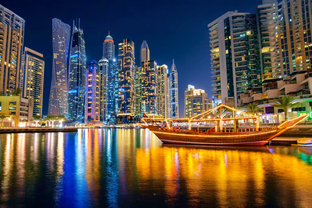 Traditional wooden dhow boat illuminated at night on Dubai Marina with glowing skyscrapers reflected in water