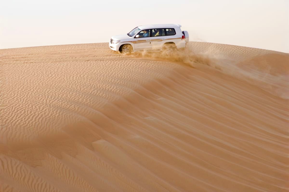 White SUV driving up sand dune in desert with dust cloud trailing behind it