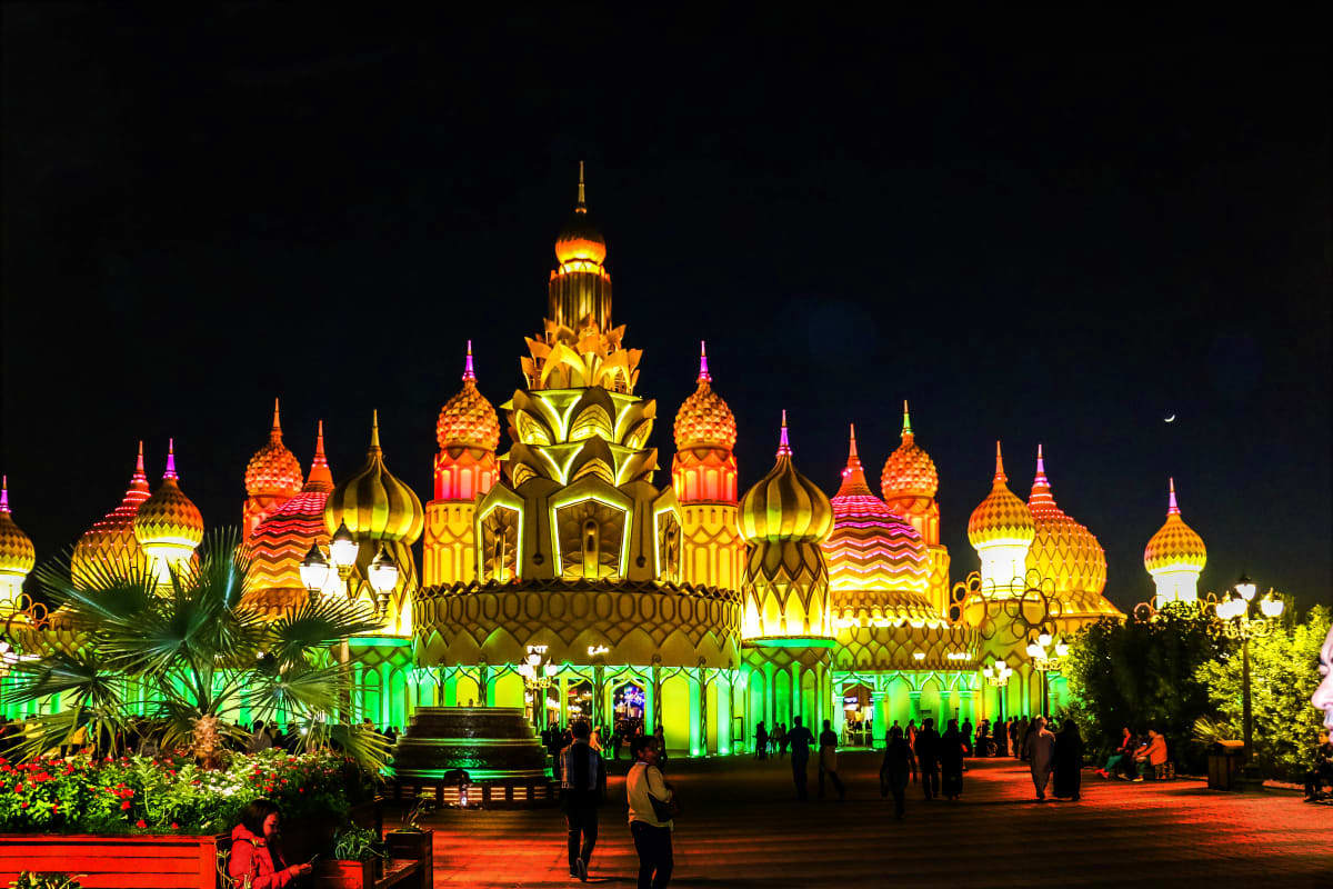 Illuminated ornate palace with golden domes and green base lit up at night with visitors walking in foreground