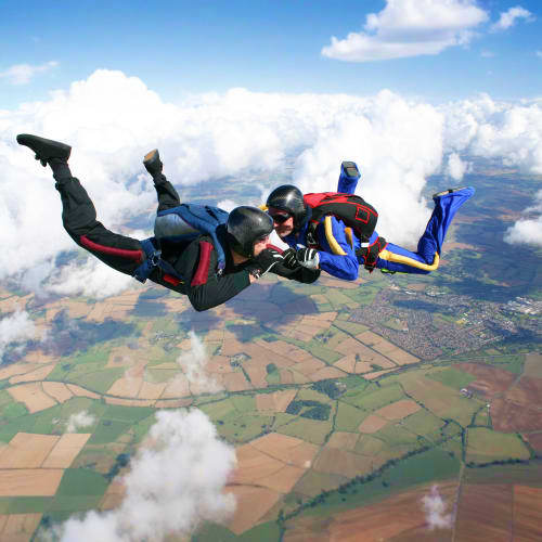 Three skydivers in formation freefalling above patchwork farmland with white clouds and blue sky