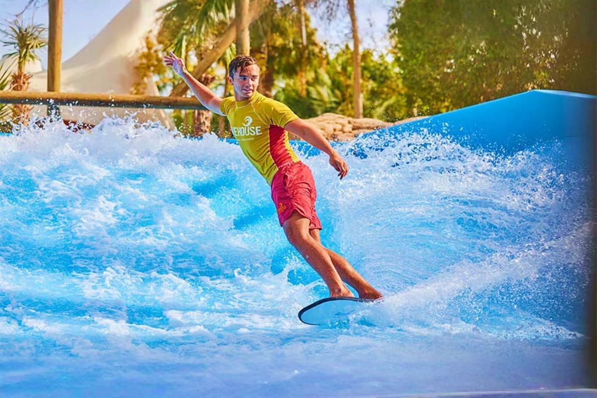 Young man surfing on artificial wave at indoor water park with palm trees in background
