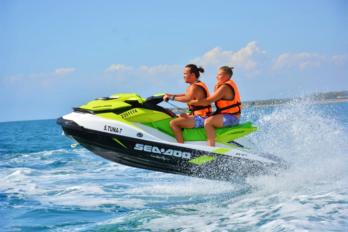 Two people riding a green and black jet ski in ocean waves near a beach coastline under blue sky