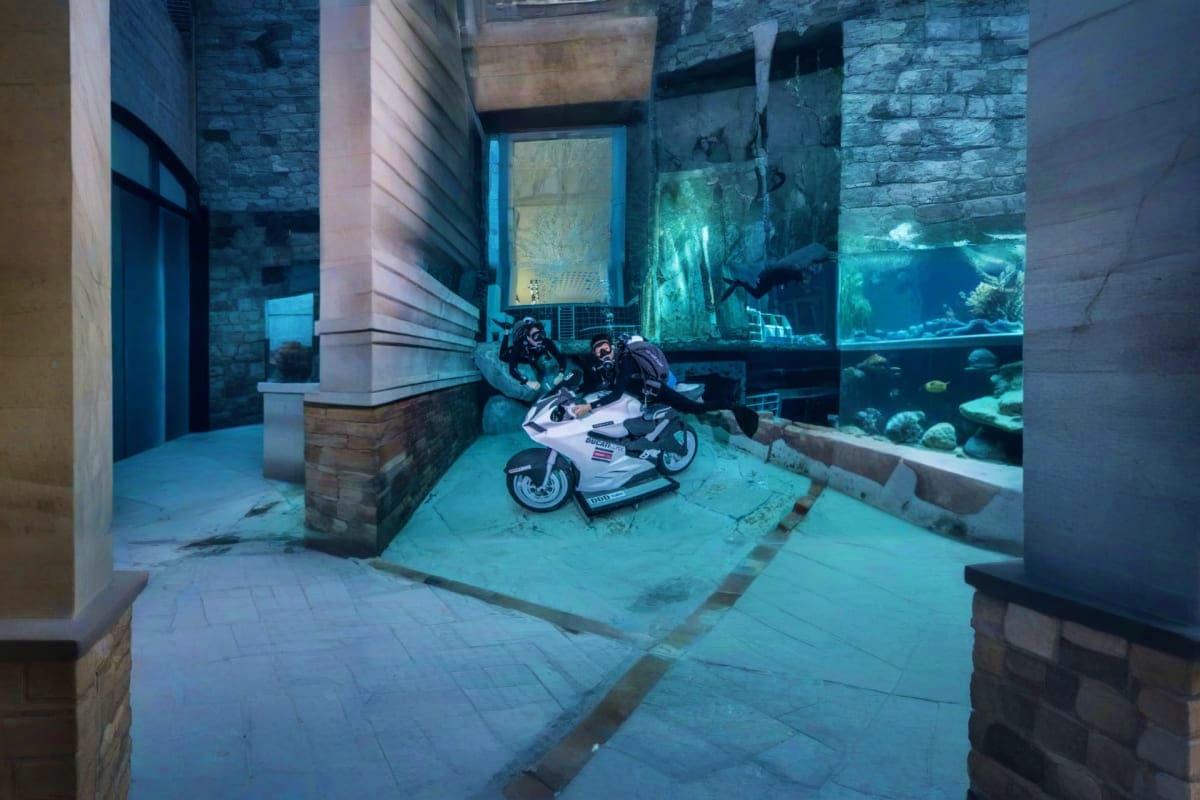 Dubai Sky Sea Adventure Escape Two scuba divers on a white and black sports motorcycle inside an underwater aquarium tunnel with fish and coral visible thro