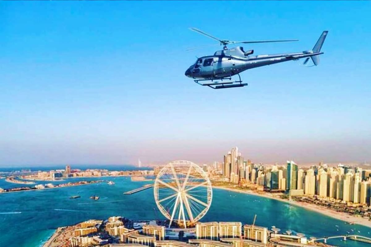 Helicopter flying over Dubai coastline with Ferris wheel, beach, and high-rise buildings visible below under clear blue sky