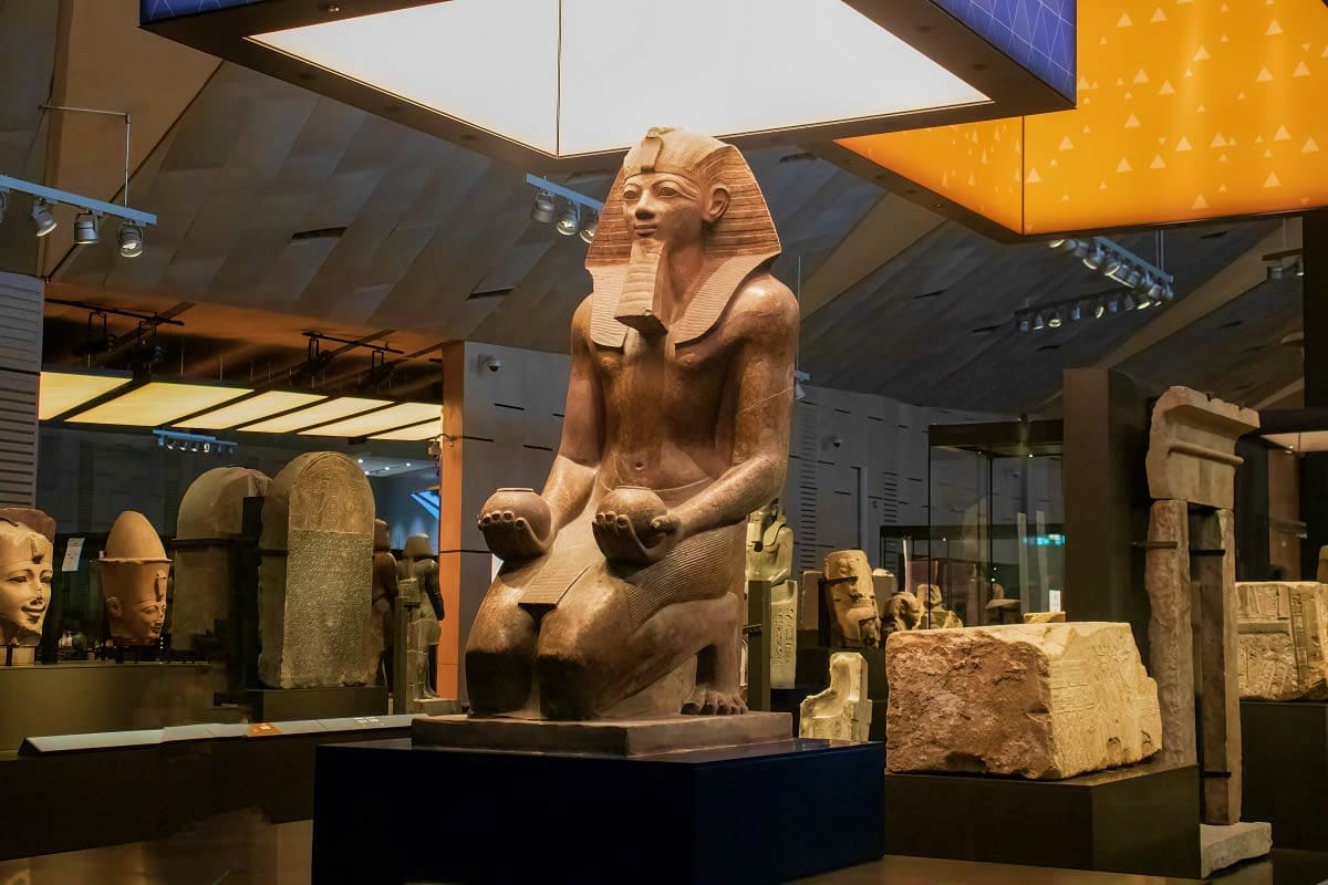 Ancient Egyptian sphinx statue on display in museum with pharaoh headdress, surrounded by other stone artifacts and sculpture