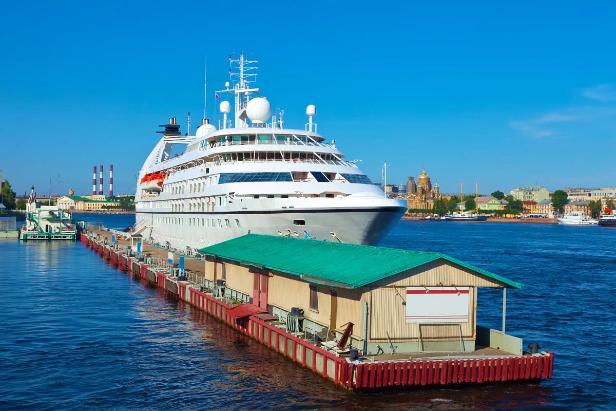 Large cruise ship docked at port with historic city landmarks visible in background under clear blue sky