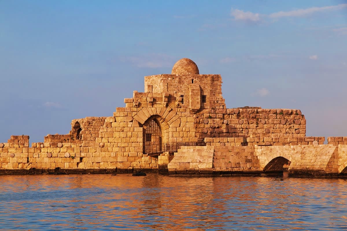 Ancient stone fortress with domed structure on waterfront, viewed from the water under clear blue sky