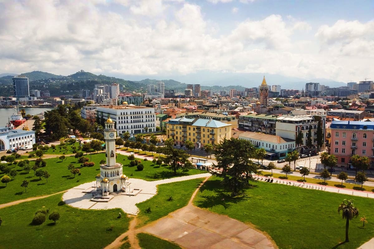 Aerial view of Batumi's seafront park with white lighthouse tower, colorful colonial buildings, and mountains under cloudy sk