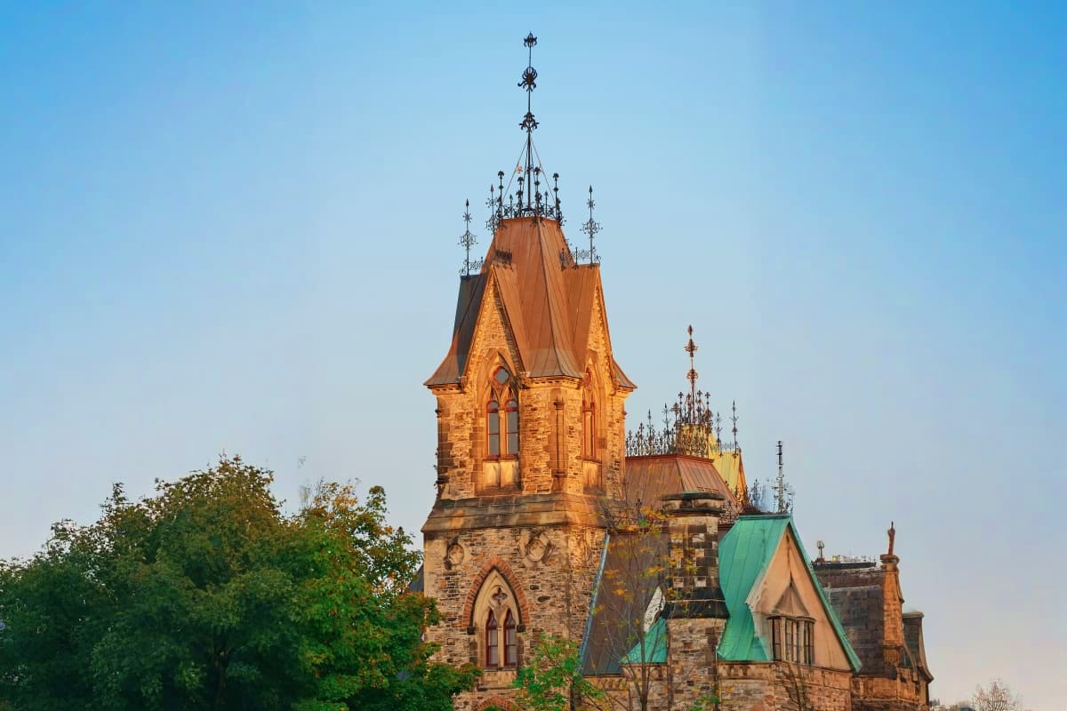 Victorian Gothic stone mansion with ornate spires and turrets against clear blue sky, surrounded by green foliage