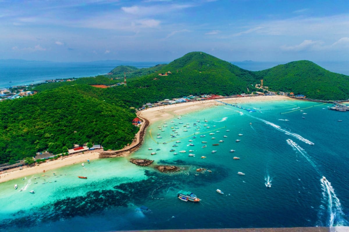 Classic Bangkok and Pattaya Holidays Aerial view of Pattaya beach with turquoise water, sandy shore, moored boats, and forested hills under blue sky