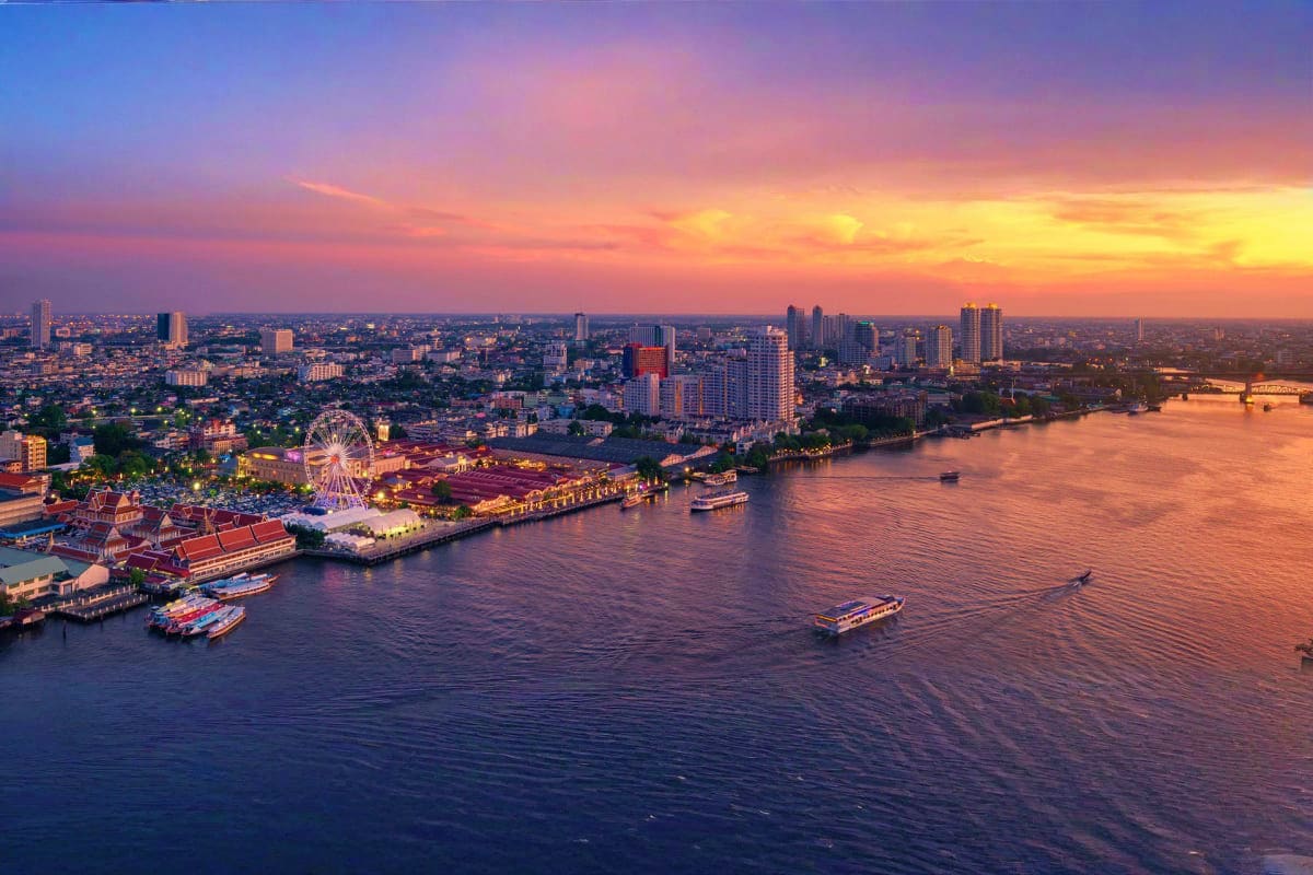 Classic Bangkok and Pattaya Holidays Aerial sunset view of Bangkok waterfront with illuminated Ferris wheel, riverside buildings, boats, and cityscape at dusk