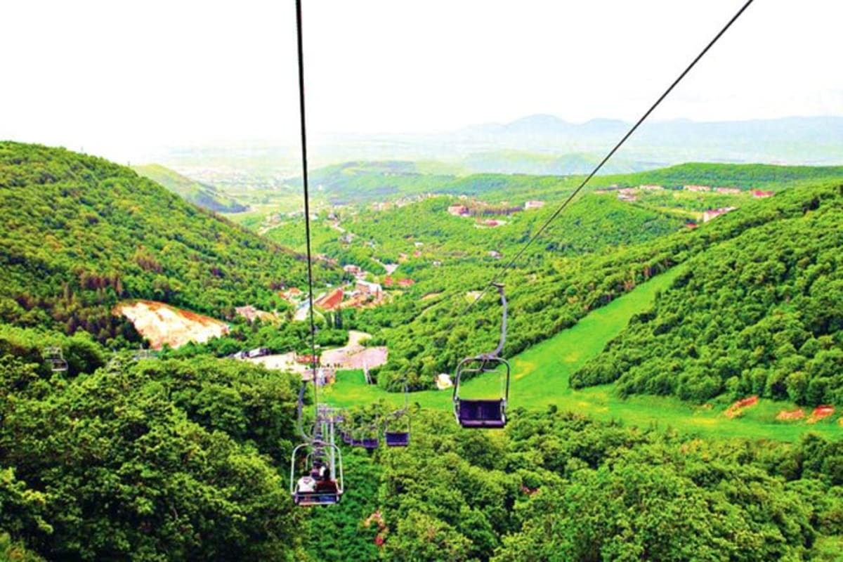Ski lift chairs ascending over lush green forested mountains and valleys with scattered villages and buildings below in Armen