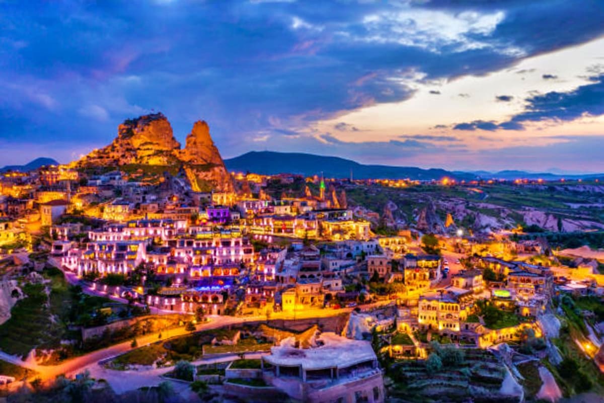 Uchisar Castle illuminated at dusk with historic stone buildings nestled in Cappadocia's fairy chimney landscape under dramat