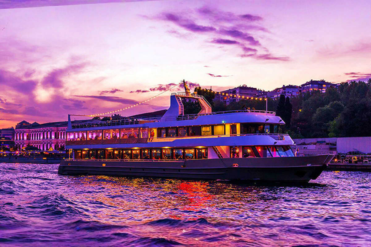 Illuminated luxury cruise ship on water at sunset with purple sky and coastal buildings in background during twilight
