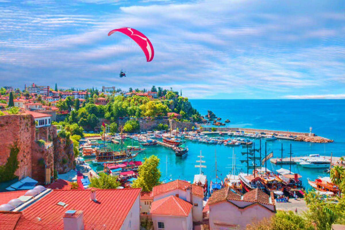 Paraglider with red parachute flying above Antalya harbor with traditional boats, red-roofed houses, and turquoise Mediterran