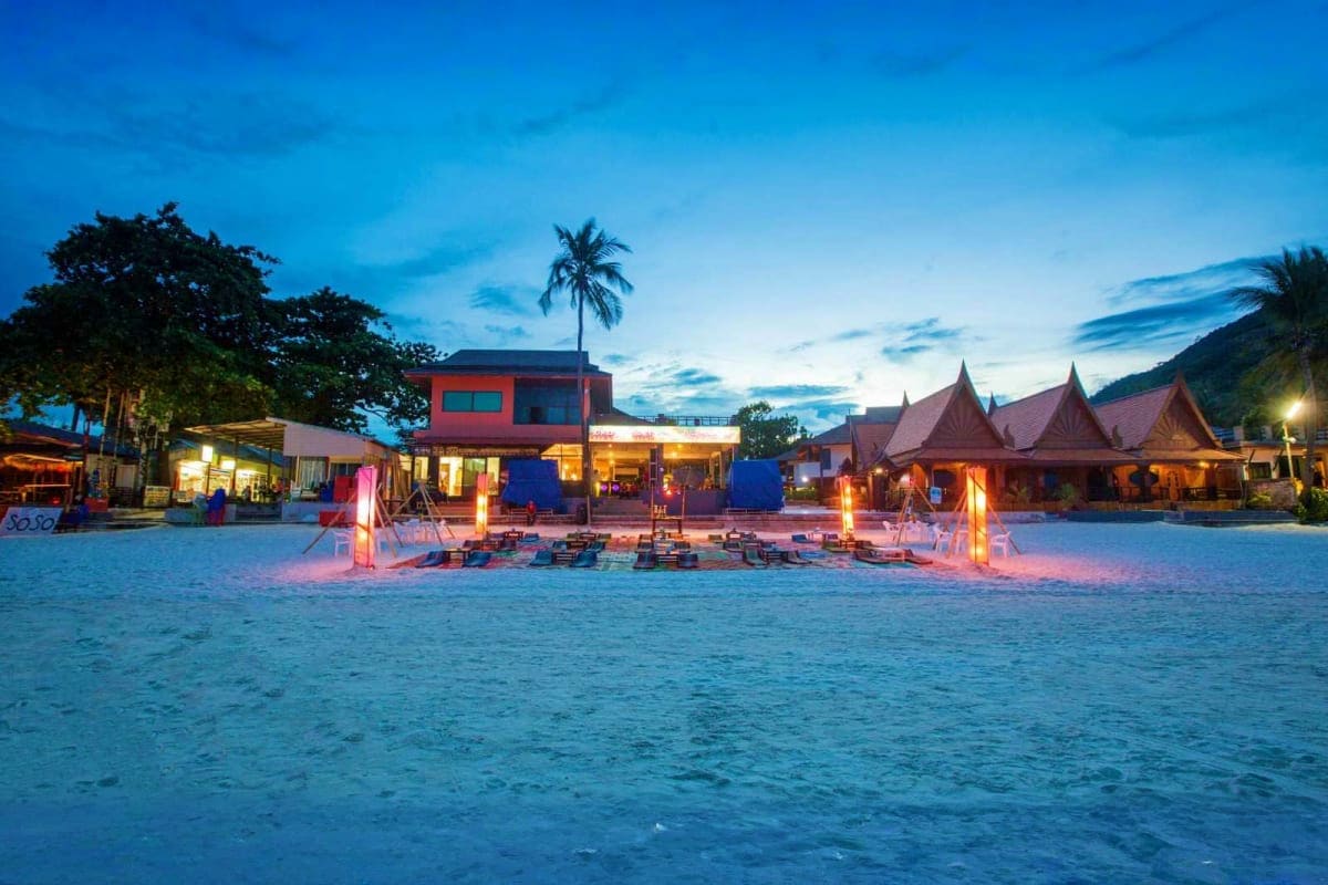 Beachfront resort at dusk with illuminated structures, beach bonfire setup, traditional Thai-style roofs, palm trees, and mou