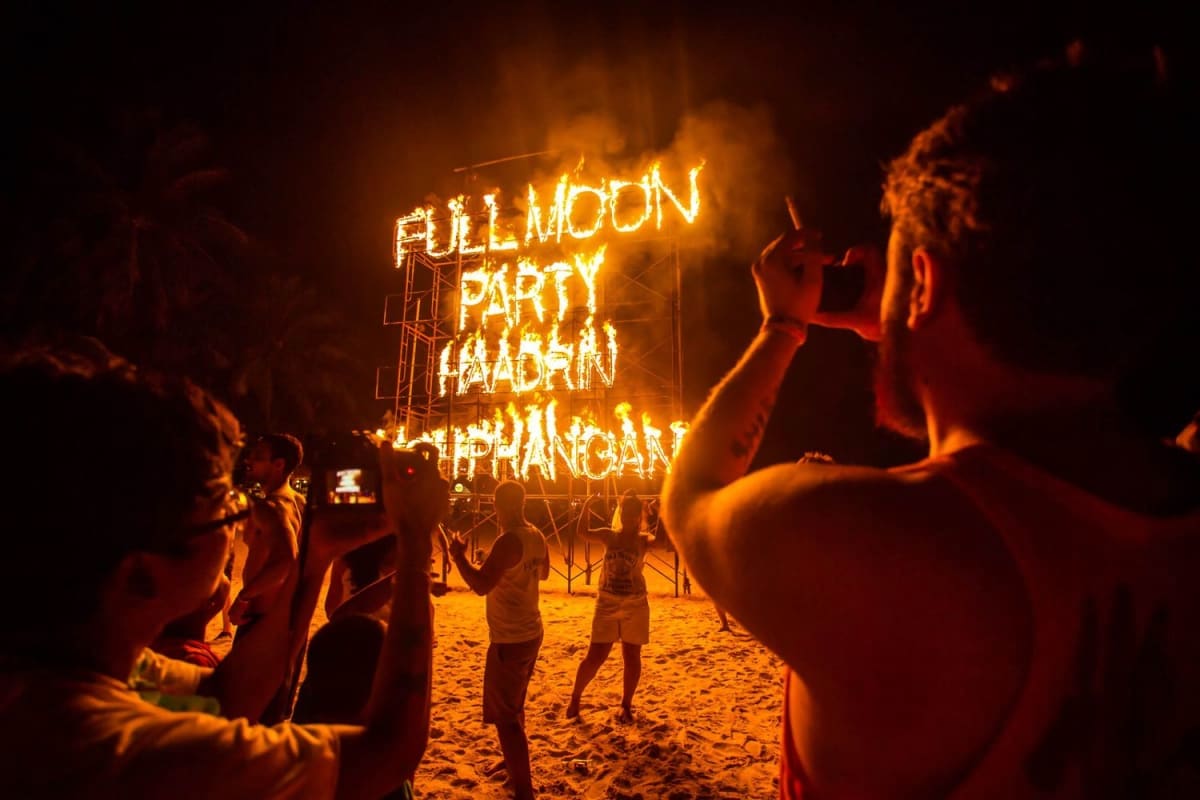 Crowded beach at night with glowing neon "Full Moon Party" sign and revelers dancing under orange lights in Koh Phangan, Thai