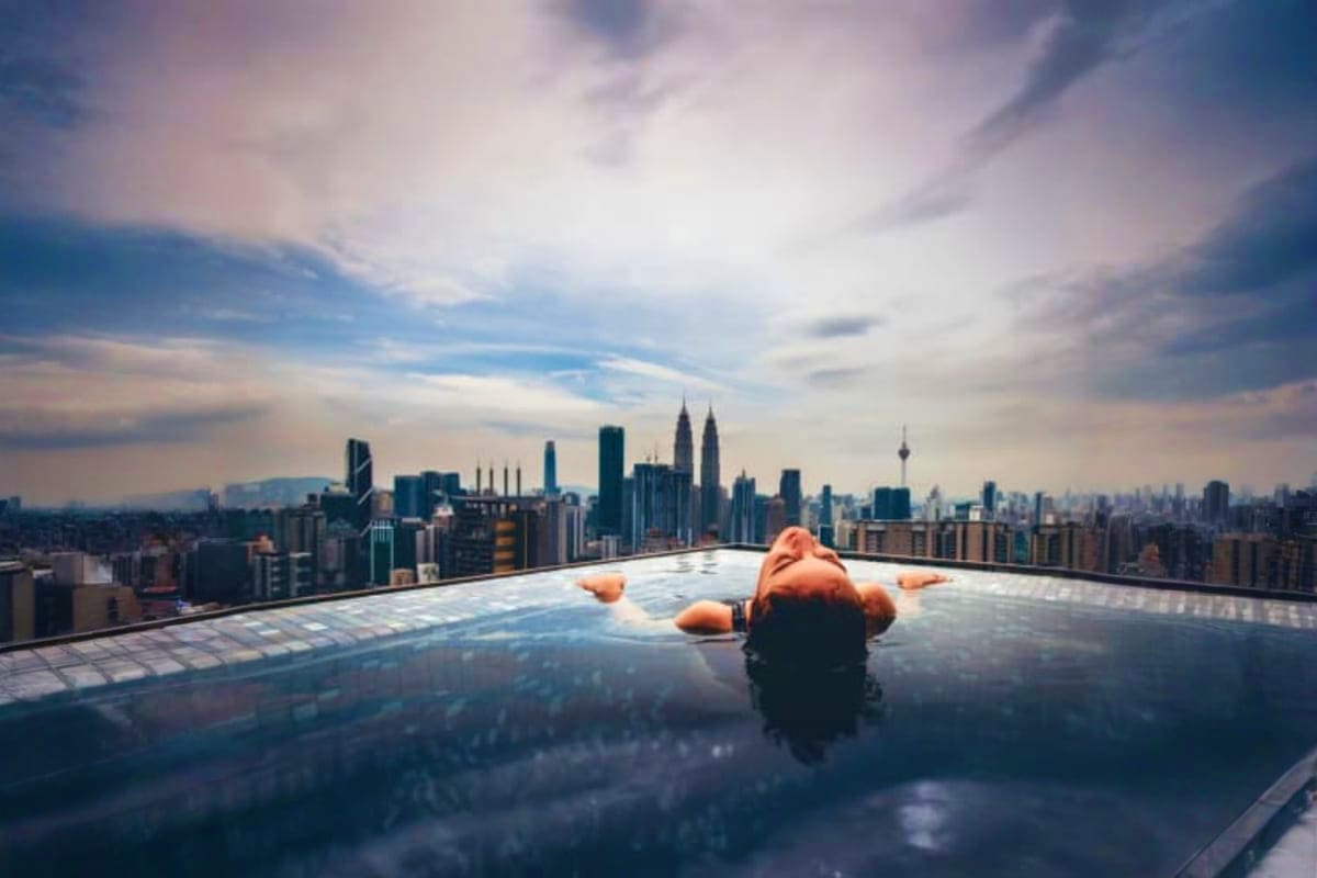 Person in orange jacket relaxing on rooftop pool overlooking Kuala Lumpur skyline with Twin Towers at sunset