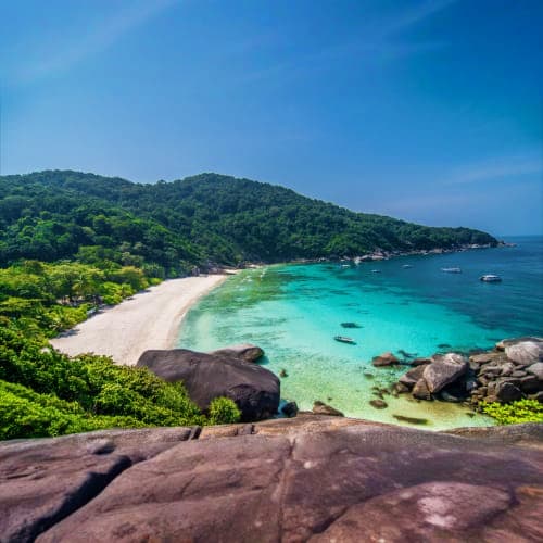 Tropical beach with turquoise waters, white sand, granite boulders, dense green hillside forest, and anchored boats under blu