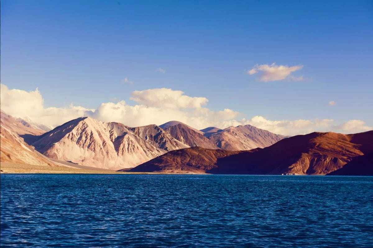Snow-capped mountains and barren peaks reflected in brilliant blue alpine lake under clear sky with white clouds