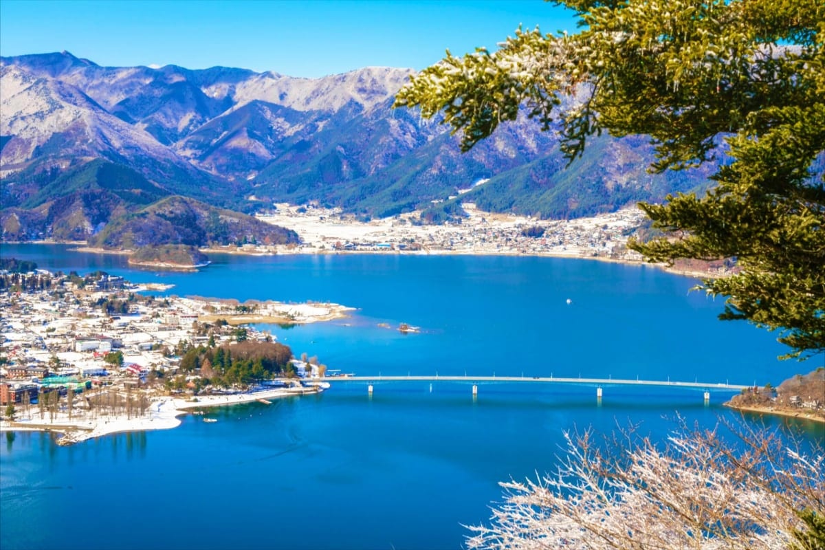 Aerial view of Lake Kawaguchi with snow-capped mountains, blue water, and a bridge connecting towns in Japan's alpine landsca