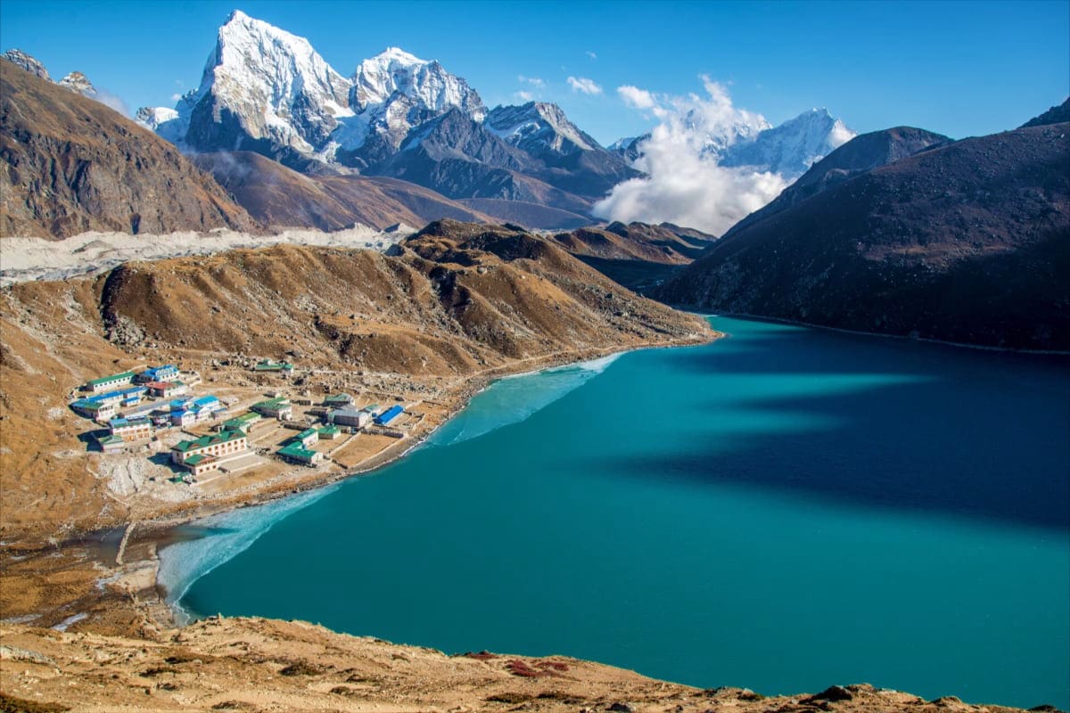 Turquoise alpine lake with snow-capped mountain peaks, brown rocky terrain, and small settlement buildings nestled on the sho