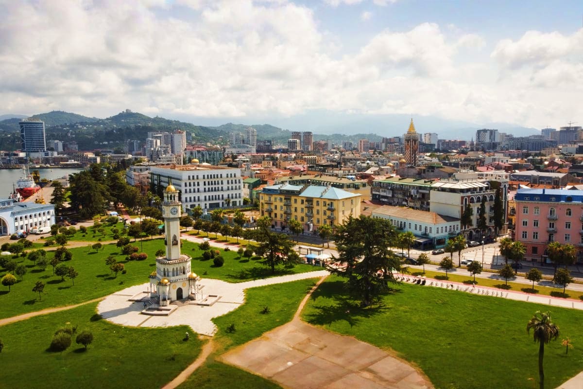 Aerial view of Tbilisi with clock tower, green park, colorful historic buildings, and mountain skyline in background