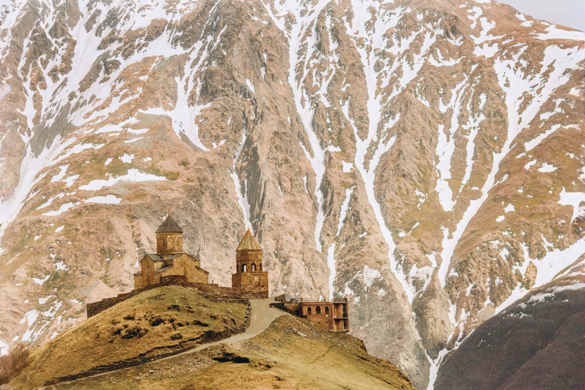 Ancient stone churches nestled on a grassy hillside with snow-capped mountain peaks and rocky terrain in the background in Ge