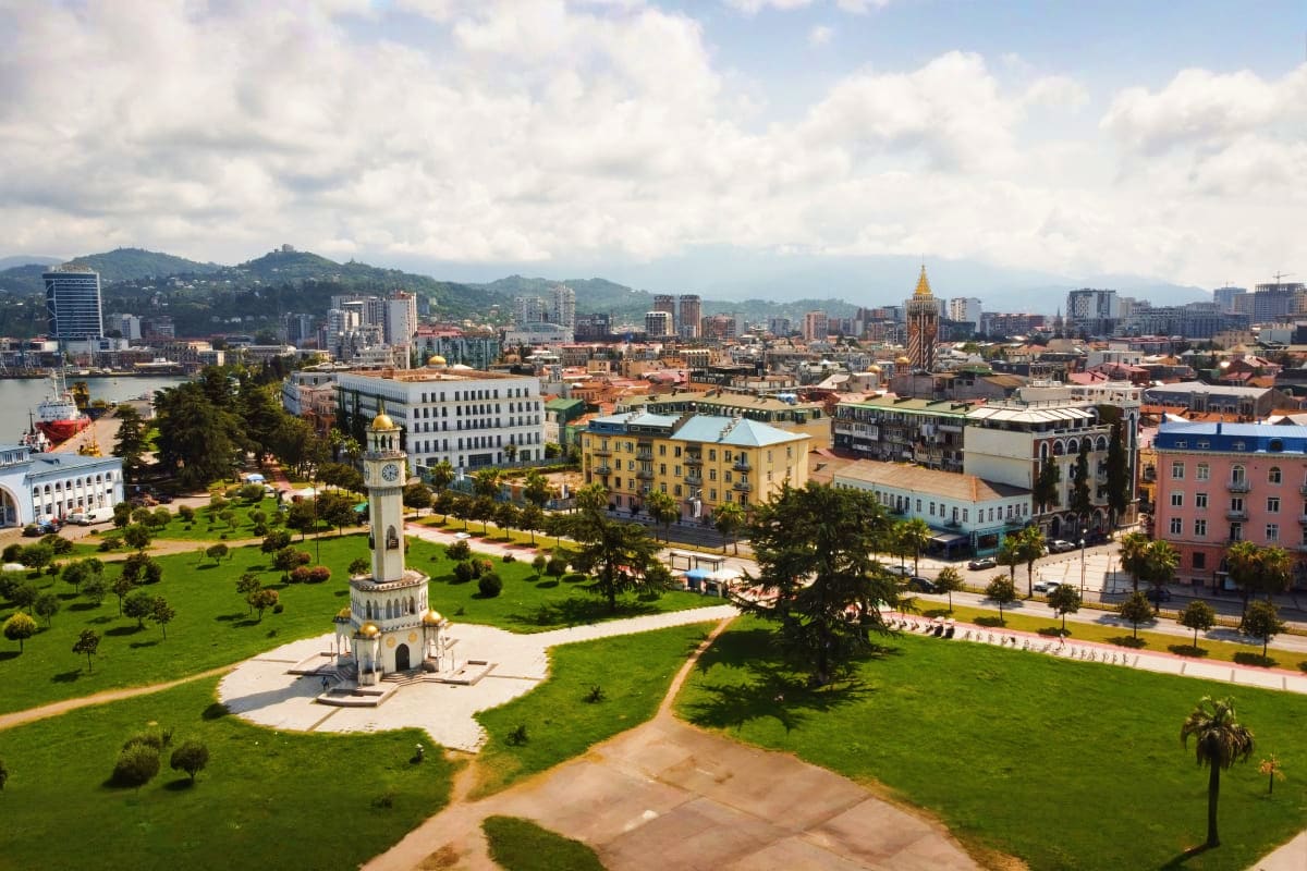 Aerial view of Tbilisi cityscape featuring white chapel tower in green park, historic buildings, and mountains under cloudy s