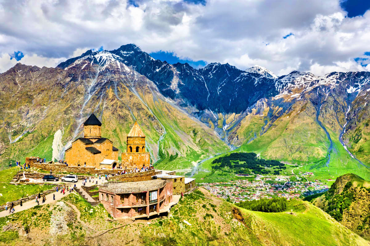 Ancient Armenian monastery with golden church and bell tower in mountainous valley with snow-capped peaks and green slopes