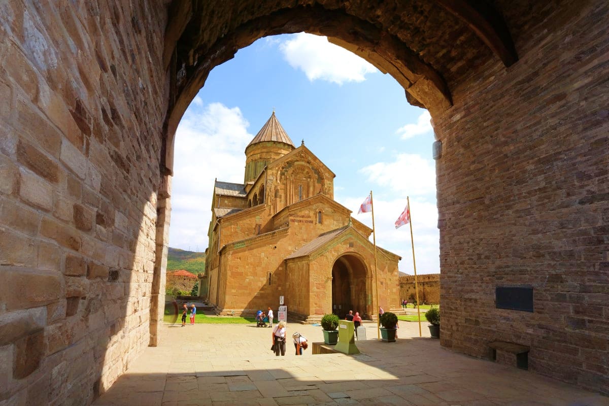 Ancient stone archway framing a golden Armenian church with conical dome and flags in a courtyard with visitors