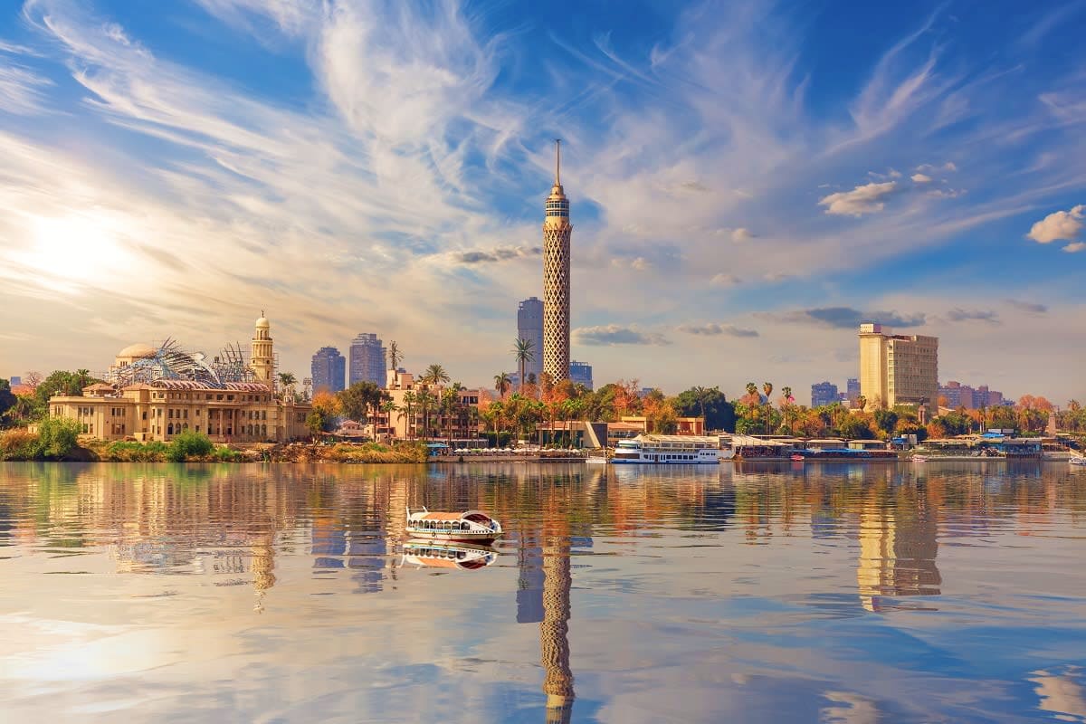 Cairo skyline with Cairo Tower and Nile River at sunrise, showing historic mosques, palm trees, and traditional felucca boat 