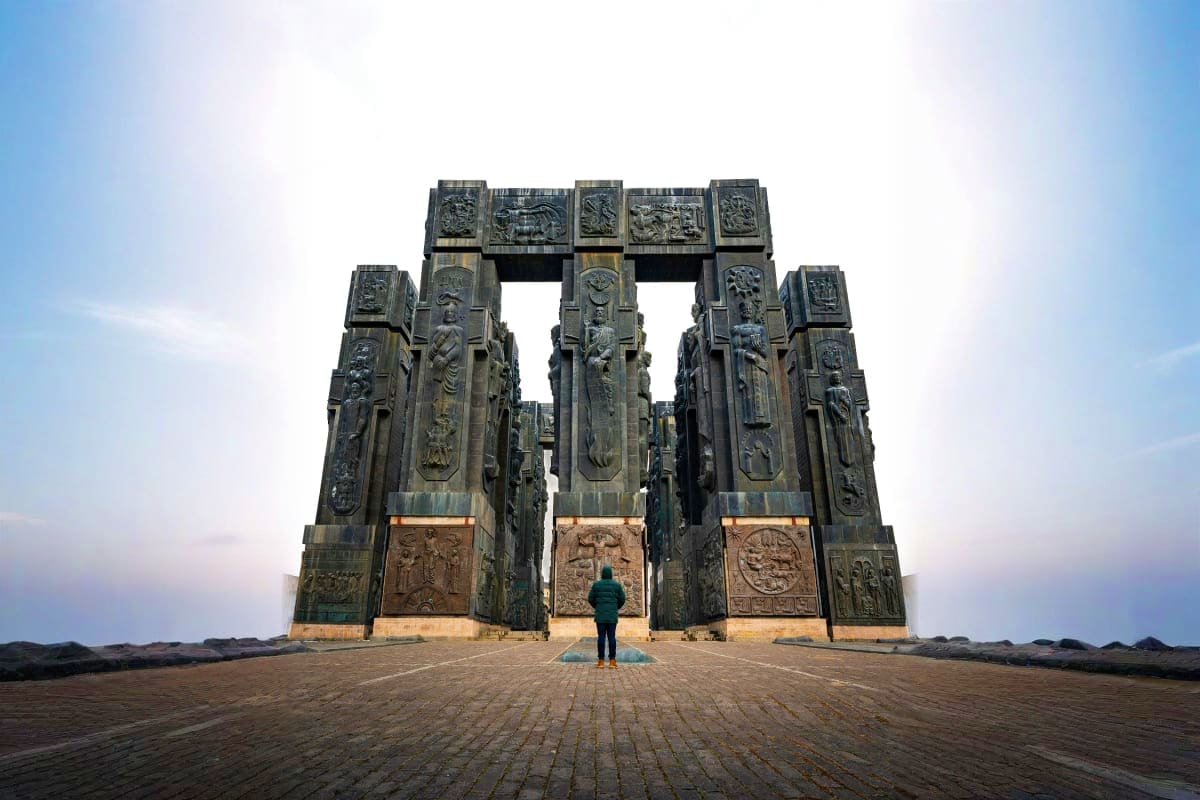 Person standing before ornate stone gateway monument with carved relief panels and symmetrical pillars under cloudy sky