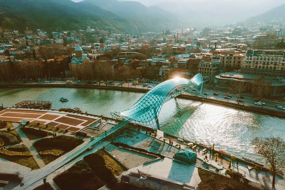 Modern glass bridge spanning river with mountain valley cityscape and traditional church in background, Tbilisi Georgia