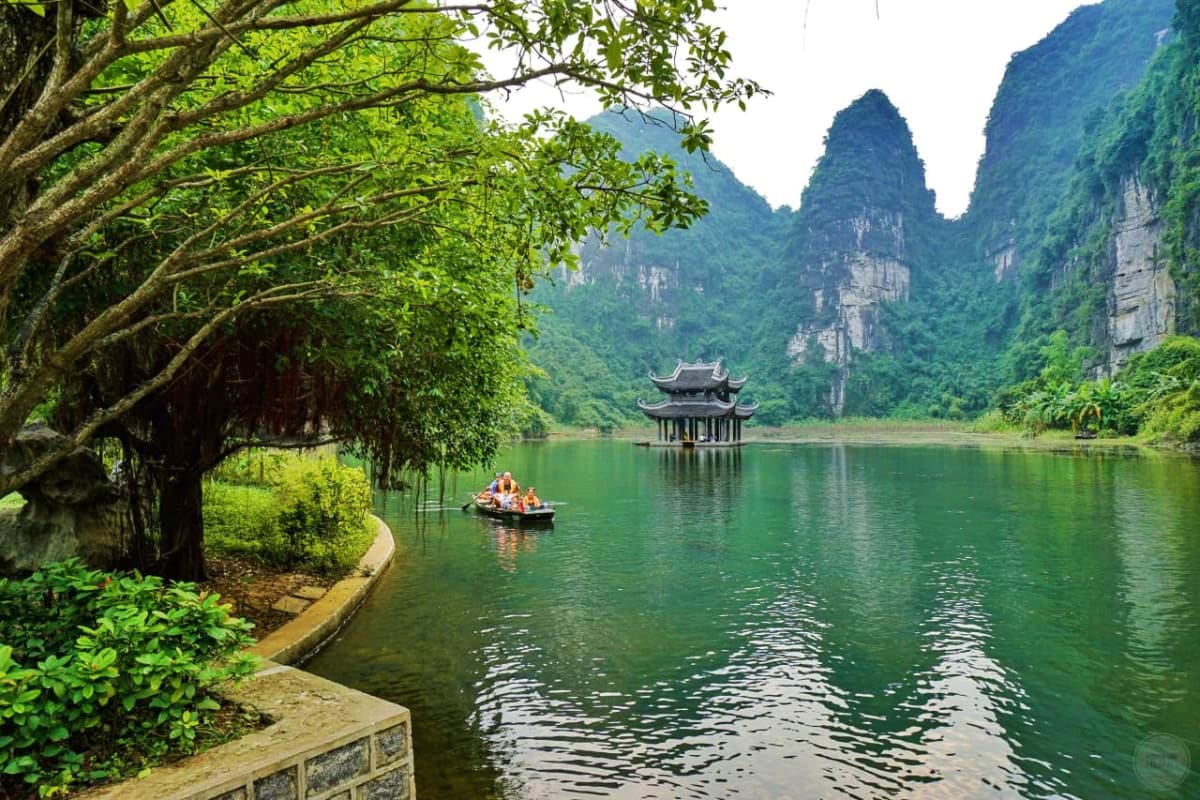 Traditional boat with tourists on emerald water beneath limestone cliffs and ancient pagoda in Trang An, Vietnam