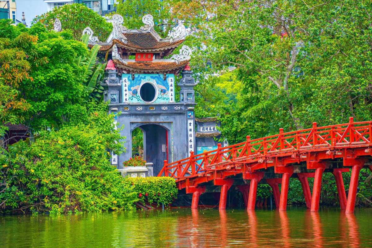 Red wooden bridge over green water leading to traditional Vietnamese pagoda with curved tile roof surrounded by lush trees