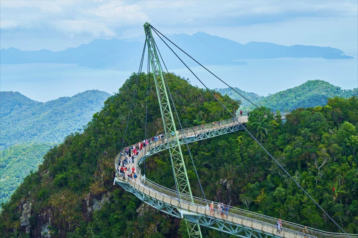 Curved suspension bridge walkway through lush forested mountains with visitors walking, overlooking misty valleys and distant