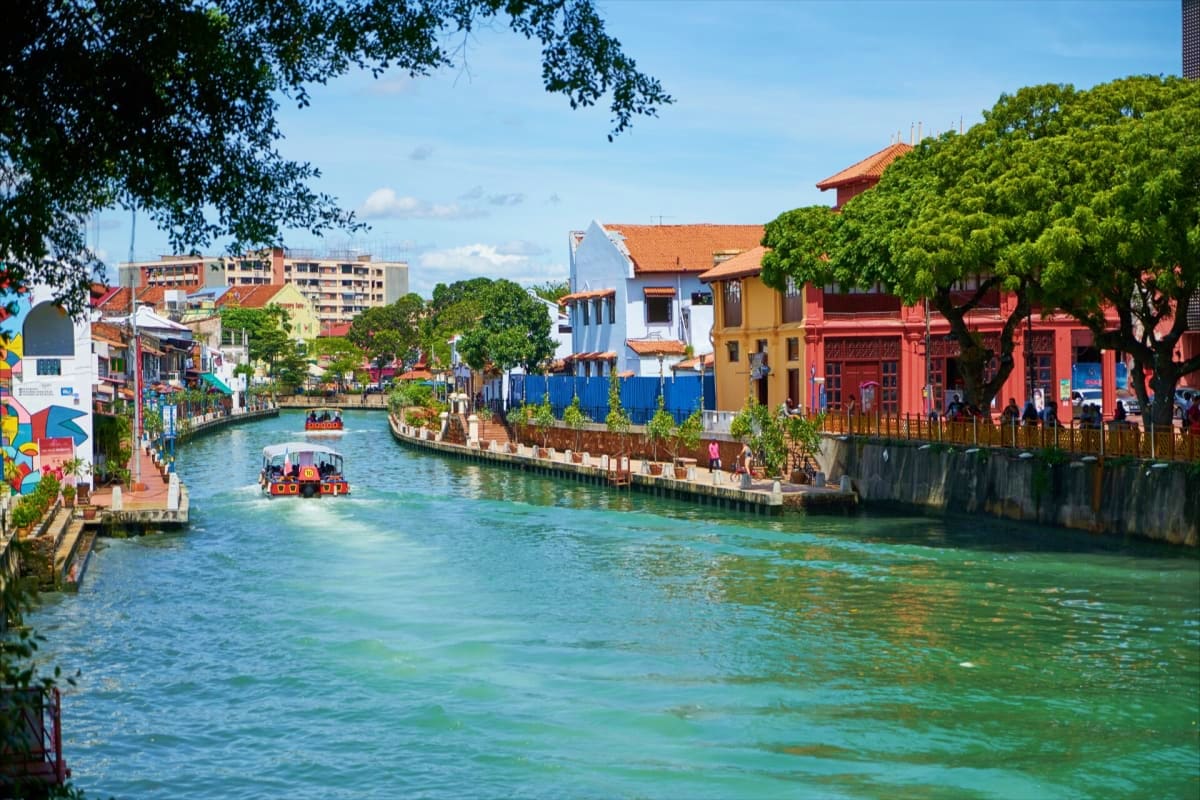 Colorful riverside buildings and tourist boats on turquoise water in Malacca, Malaysia's historic riverside district.