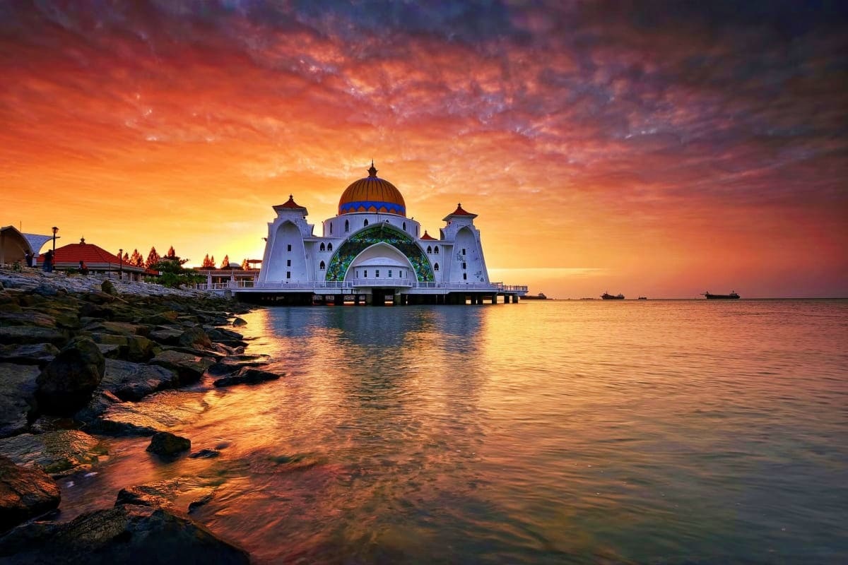 Masjid Selat mosque with blue dome silhouetted at sunset over calm sea waters with rocky shore in foreground