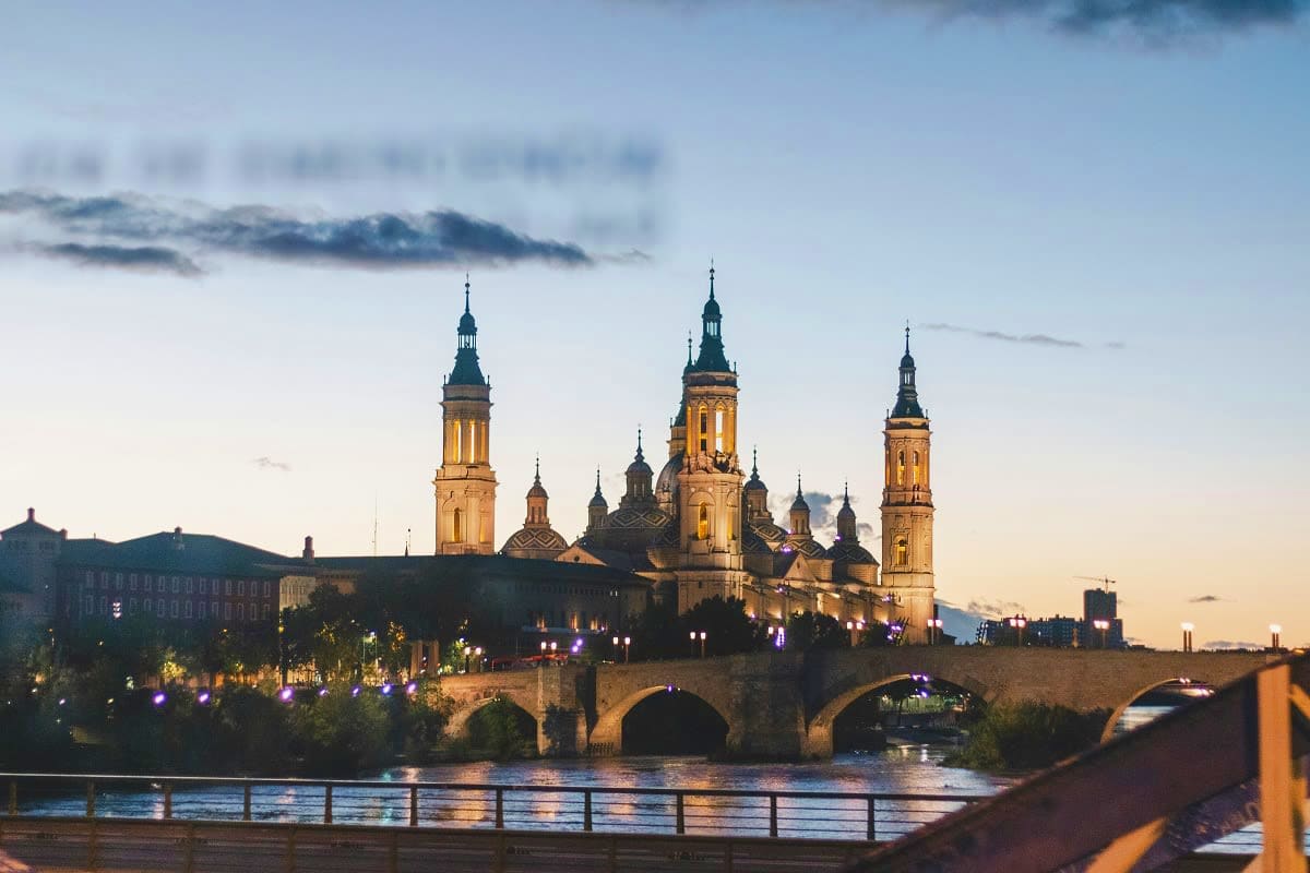 Basilica of Our Lady of the Pillar illuminated at dusk with stone bridge crossing Ebro River in Zaragoza, Spain