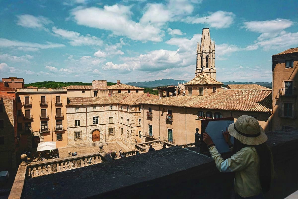 Tourist photographing historic Spanish town square with church bell tower and terracotta-roofed buildings under blue sky