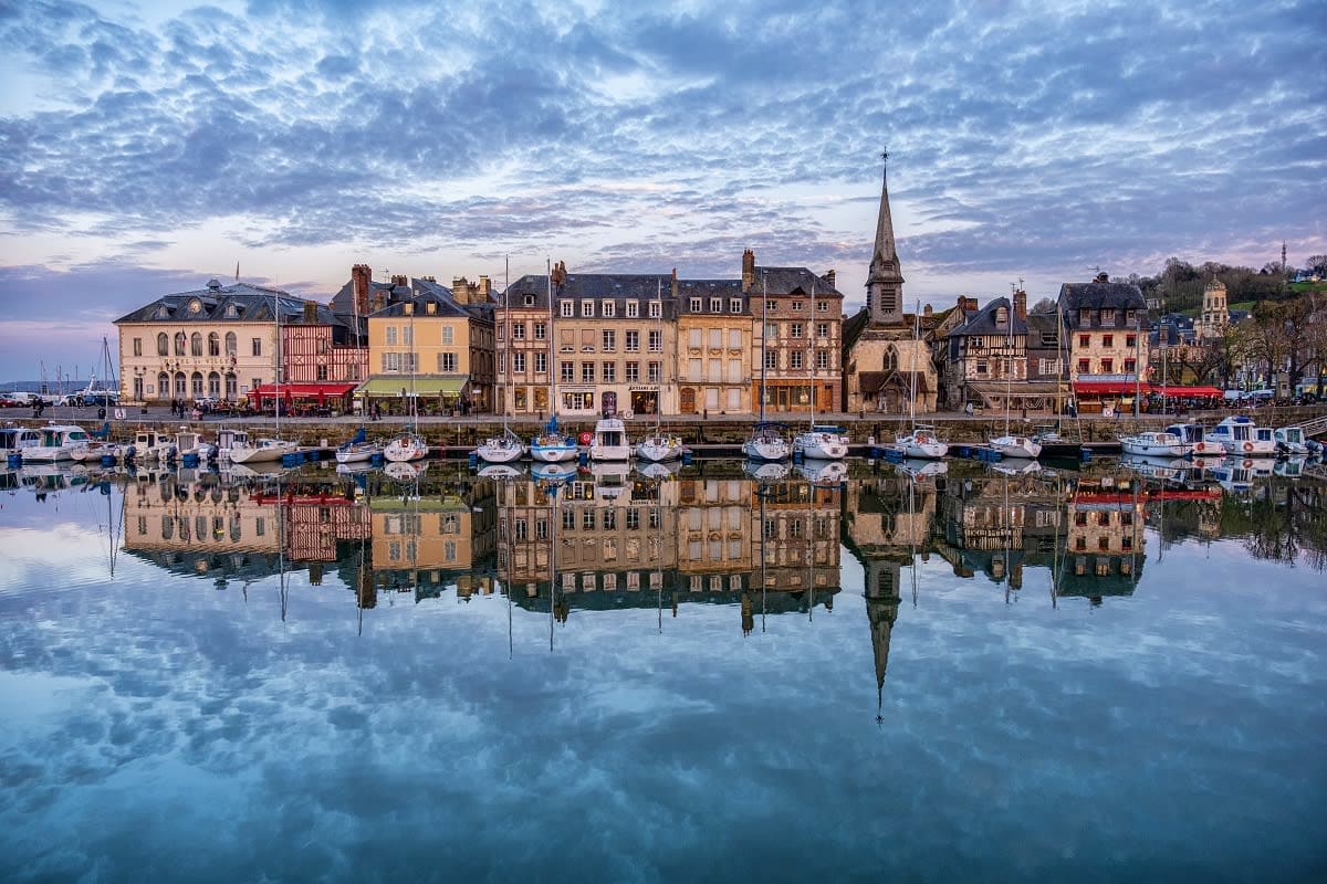 Historic European harbor town with colorful waterfront buildings, moored sailboats, and church spire reflected in calm water 