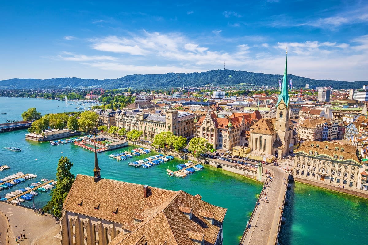 Aerial view of Zurich's old town with turquoise Limmat River, historic churches, boats, and forested hills in background