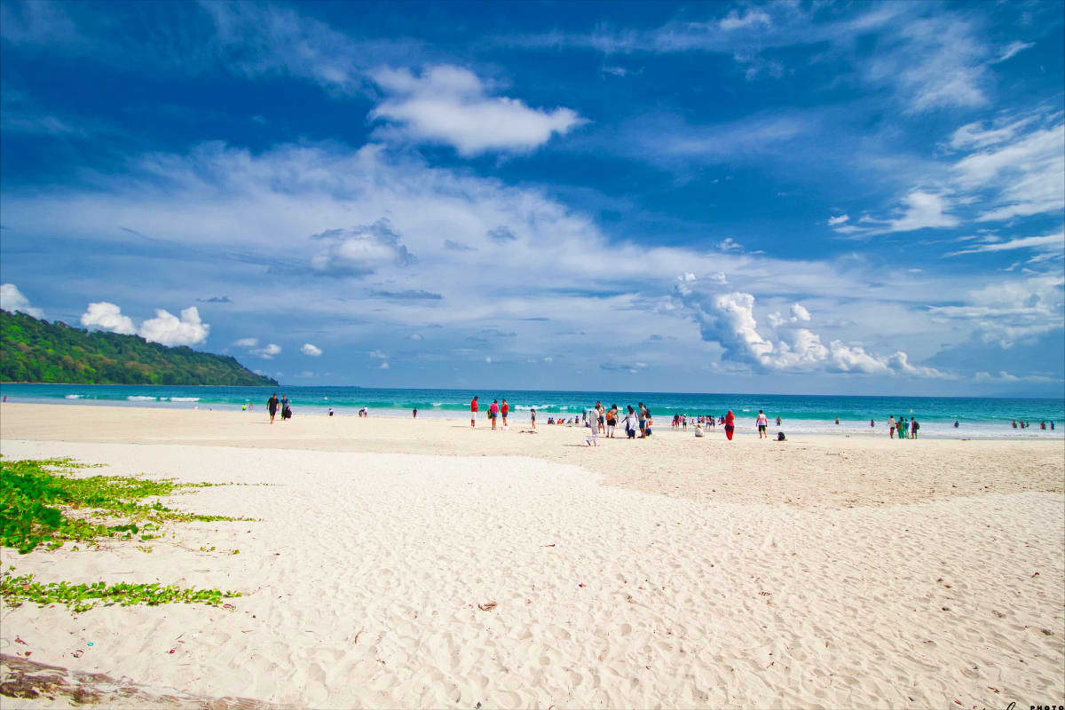 Crowded tropical beach with white sand, turquoise water, and forested hillside under blue sky with white clouds